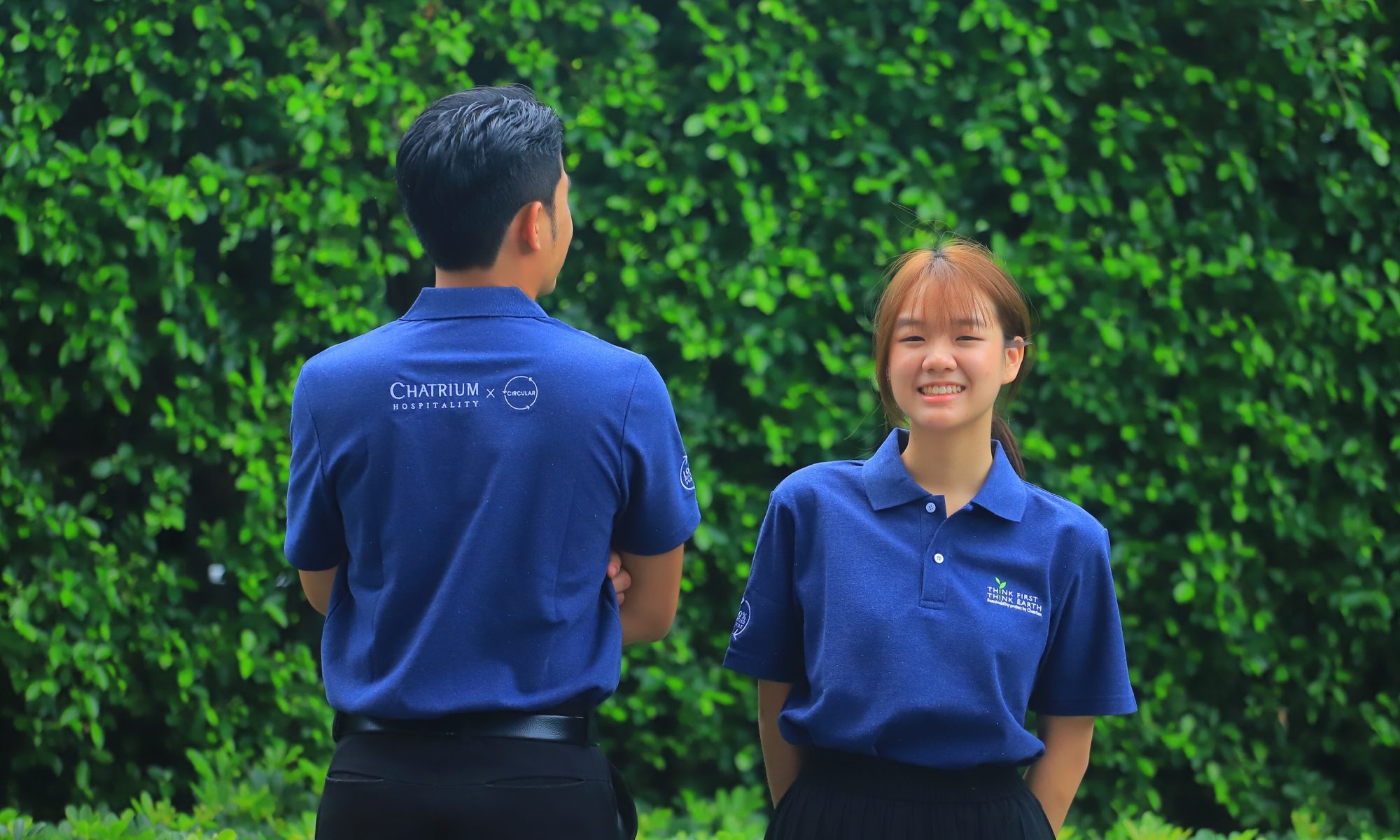 Two individuals wearing blue shirts posing in front of bushes.