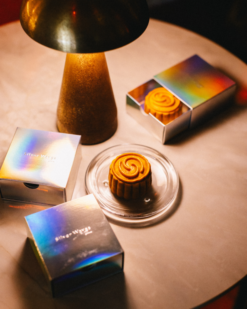 Individual mooncakes from Silver Waves by Boon are presented in iridescent gift boxes on a marble table under warm ambient lighting. One mooncake is displayed on a clear glass plate while another sits in an open box, showcasing the elegant single serving packaging and handcrafted design.