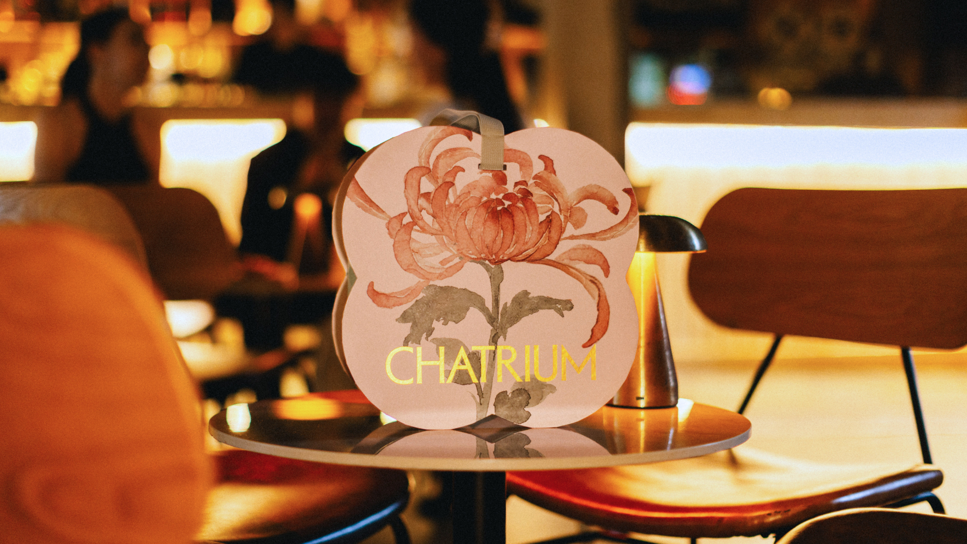 A Chatrium Mooncake gift box featuring a hand painted chrysanthemum illustration and gold Chatrium logo sits on a small round table in a softly lit restaurant. The warm, blurred background creates an inviting atmosphere, highlighting the elegant seasonal packaging inspired by the Mid Autumn Festival.